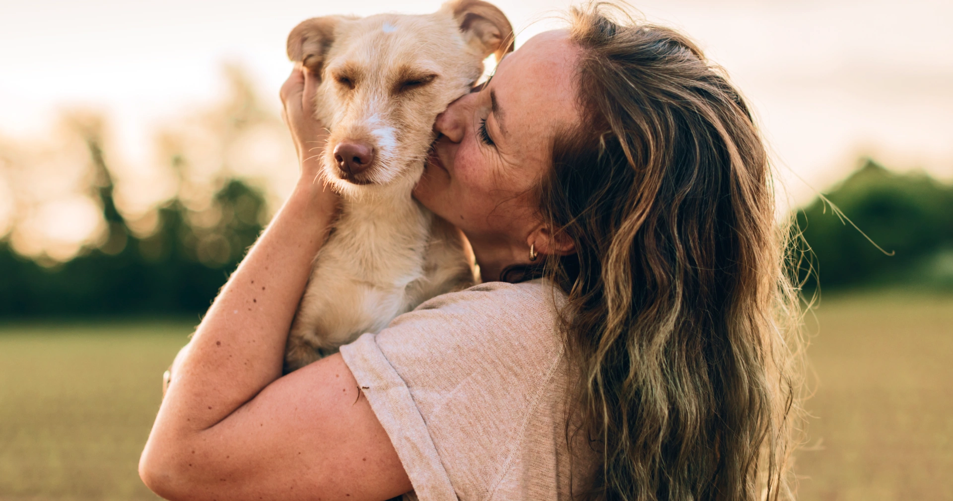 A woman with long wavy hair holding and kissing a light-colored dog outdoors in a field during warm golden hour light.